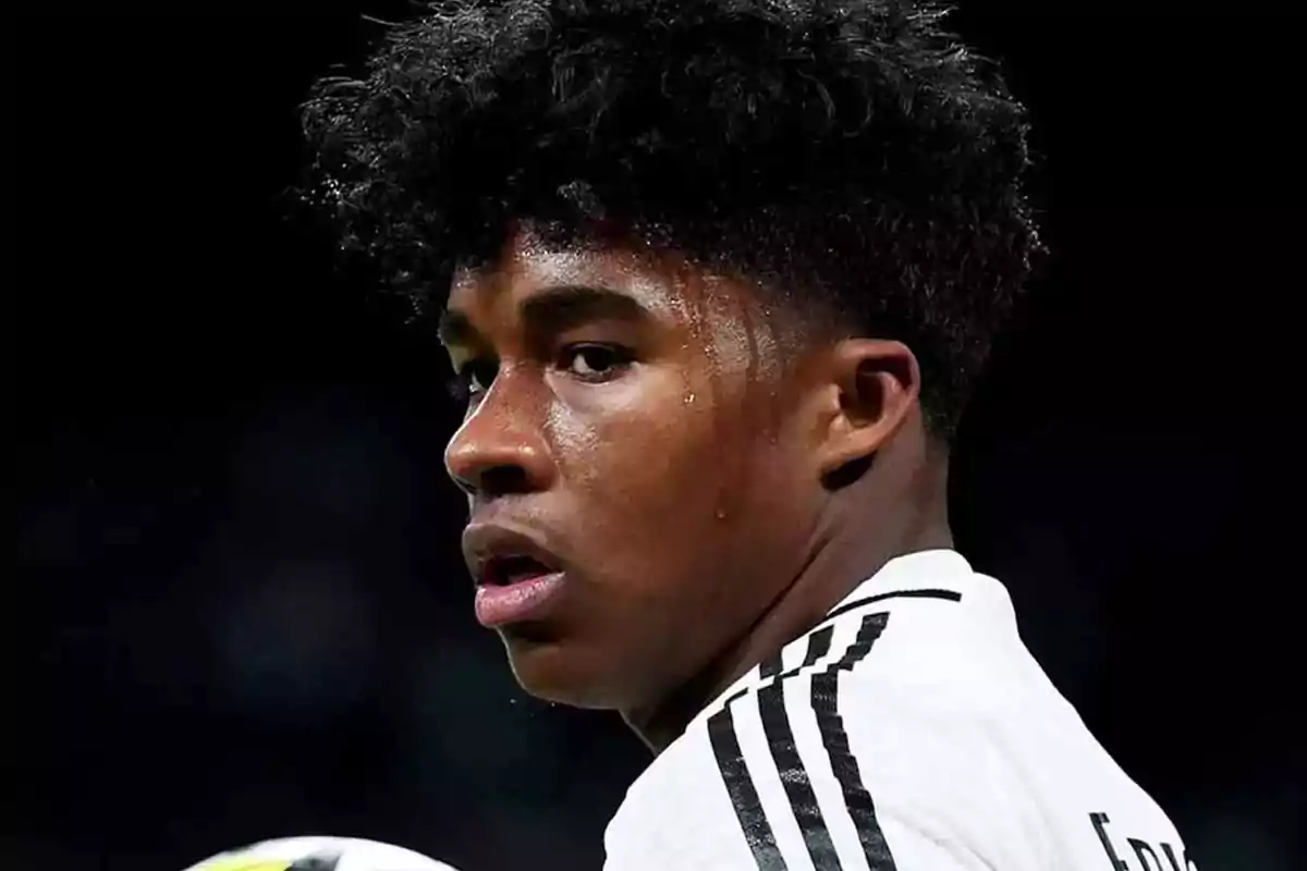 Un jugador de fútbol con camiseta blanca y rayas negras mira hacia un lado en un campo oscuro. Un jugador de fútbol con camiseta blanca y rayas negras mira hacia un lado en un campo oscuro.