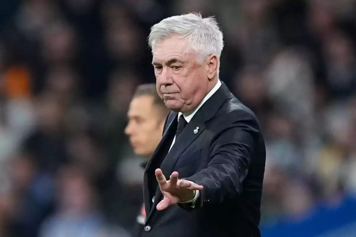 A gray-haired man in a dark suit gestures with his hand in a sports setting.