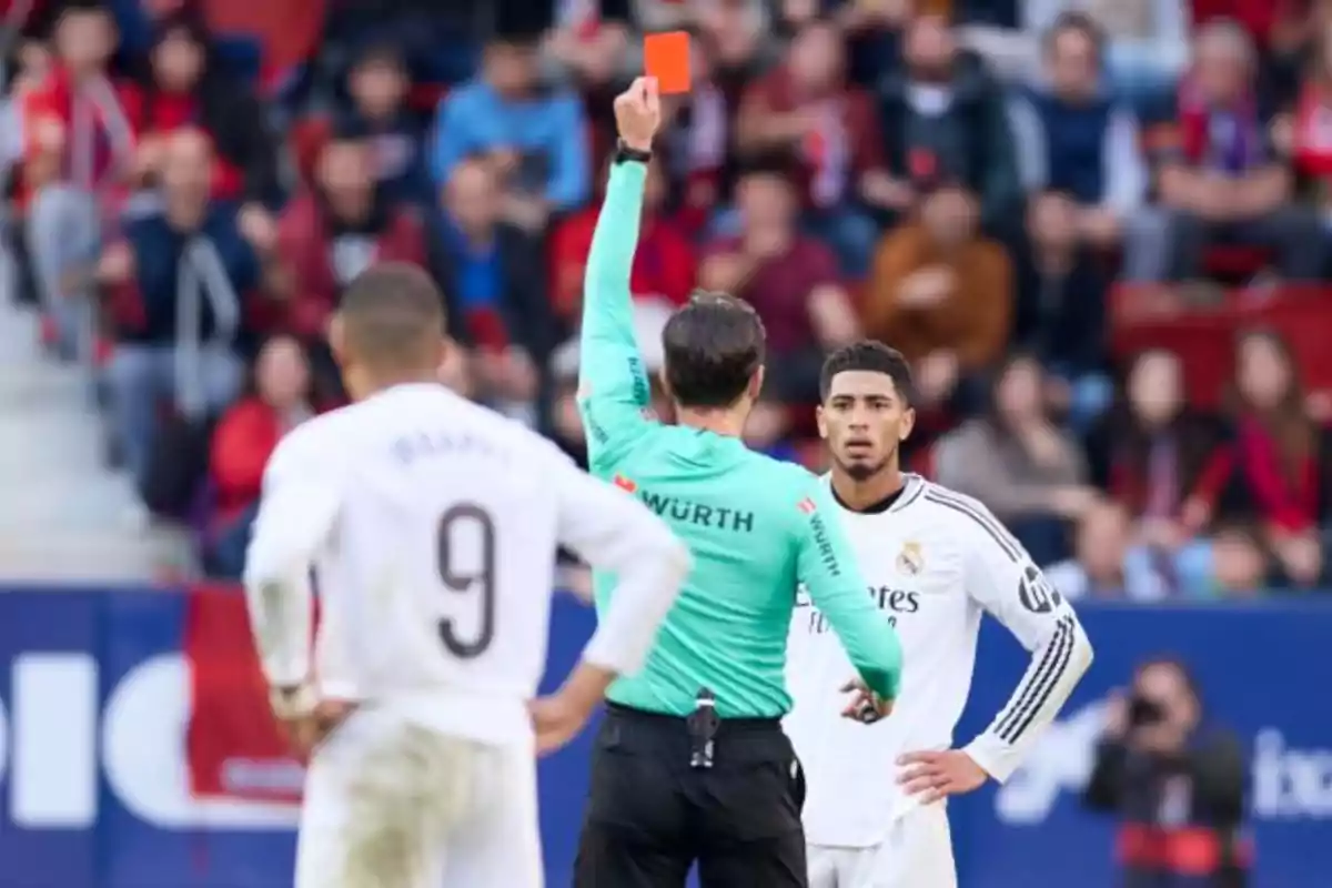 Un árbitro muestra una tarjeta roja a un jugador de fútbol durante un partido, mientras otro jugador observa la escena.