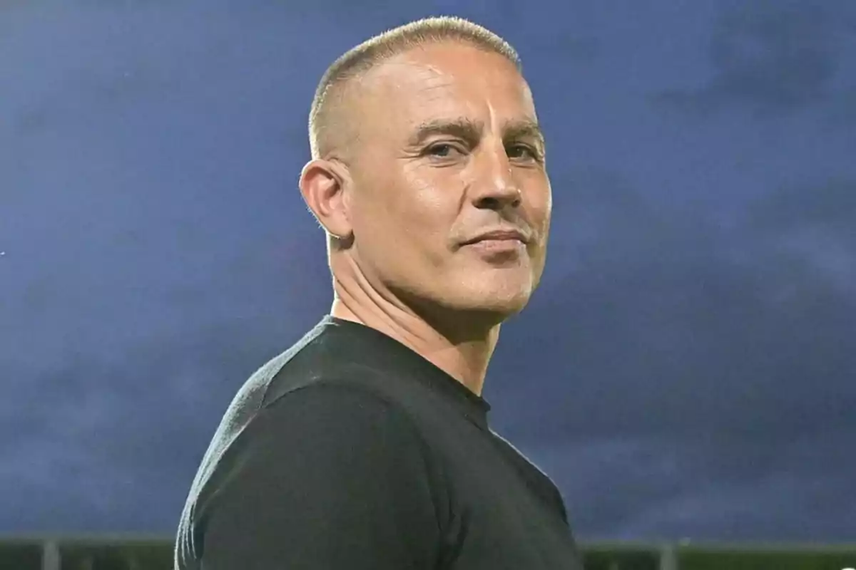 A man with short hair and a black t-jersey looks toward the camera with a blue sky in the background. A man with short hair and a black t-jersey looks toward the camera with a blue sky in the background.