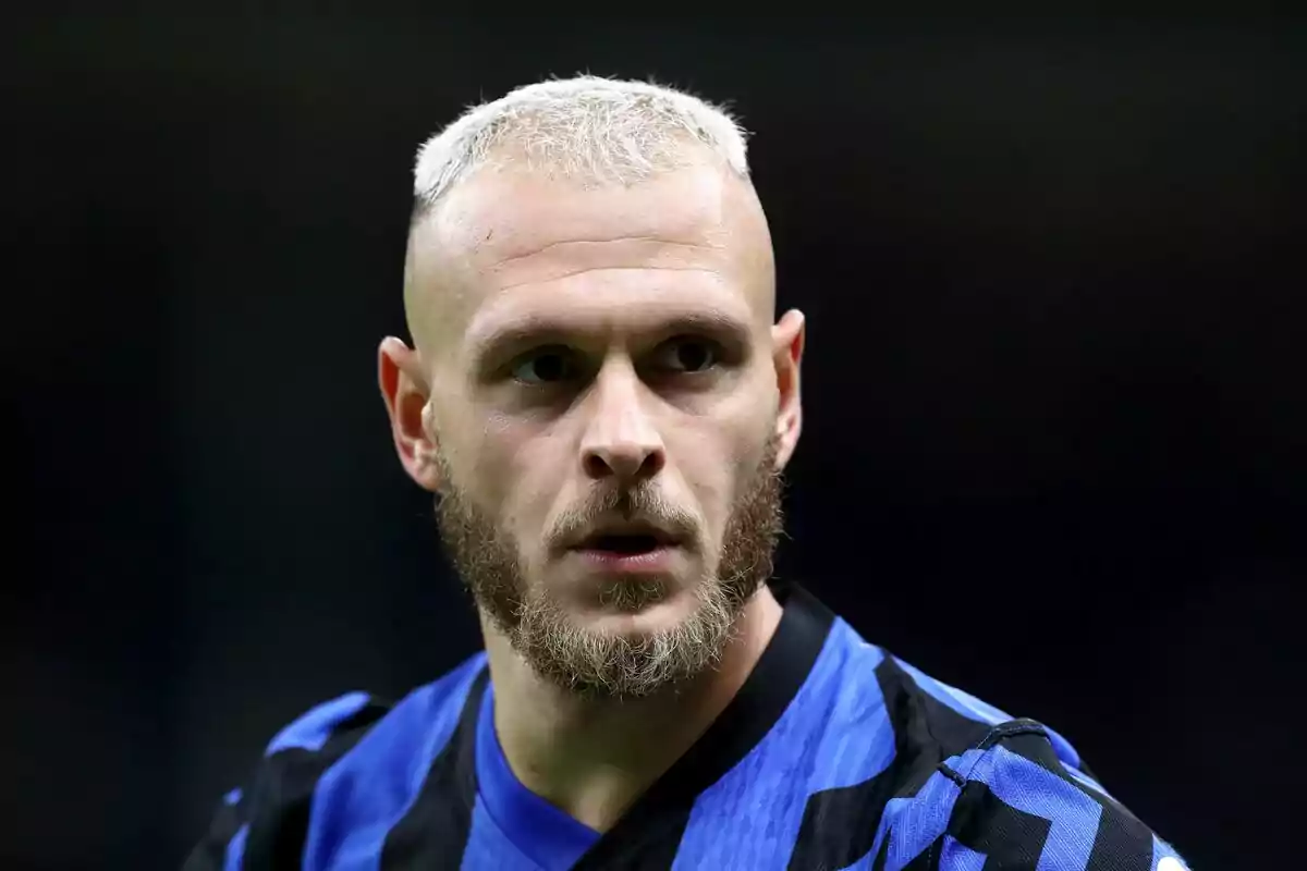 A player with blonde hair and a beard, wearing a blue and black jersey, looks to the side in a stadium.