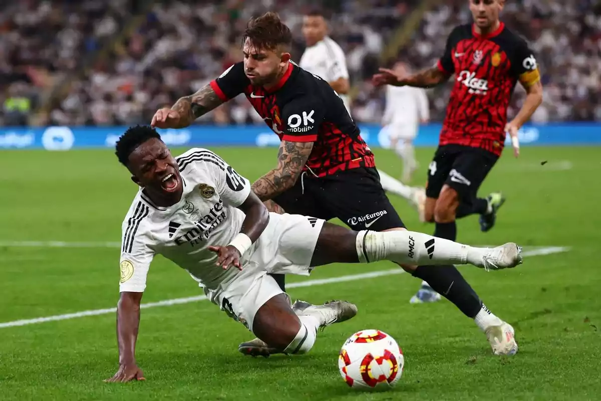 Un jugador de fútbol con uniforme blanco cae al suelo mientras otro jugador con uniforme negro y rojo lo desafía en un partido. Un jugador de fútbol con uniforme blanco cae al suelo mientras otro jugador con uniforme negro y rojo lo desafía en un partido.