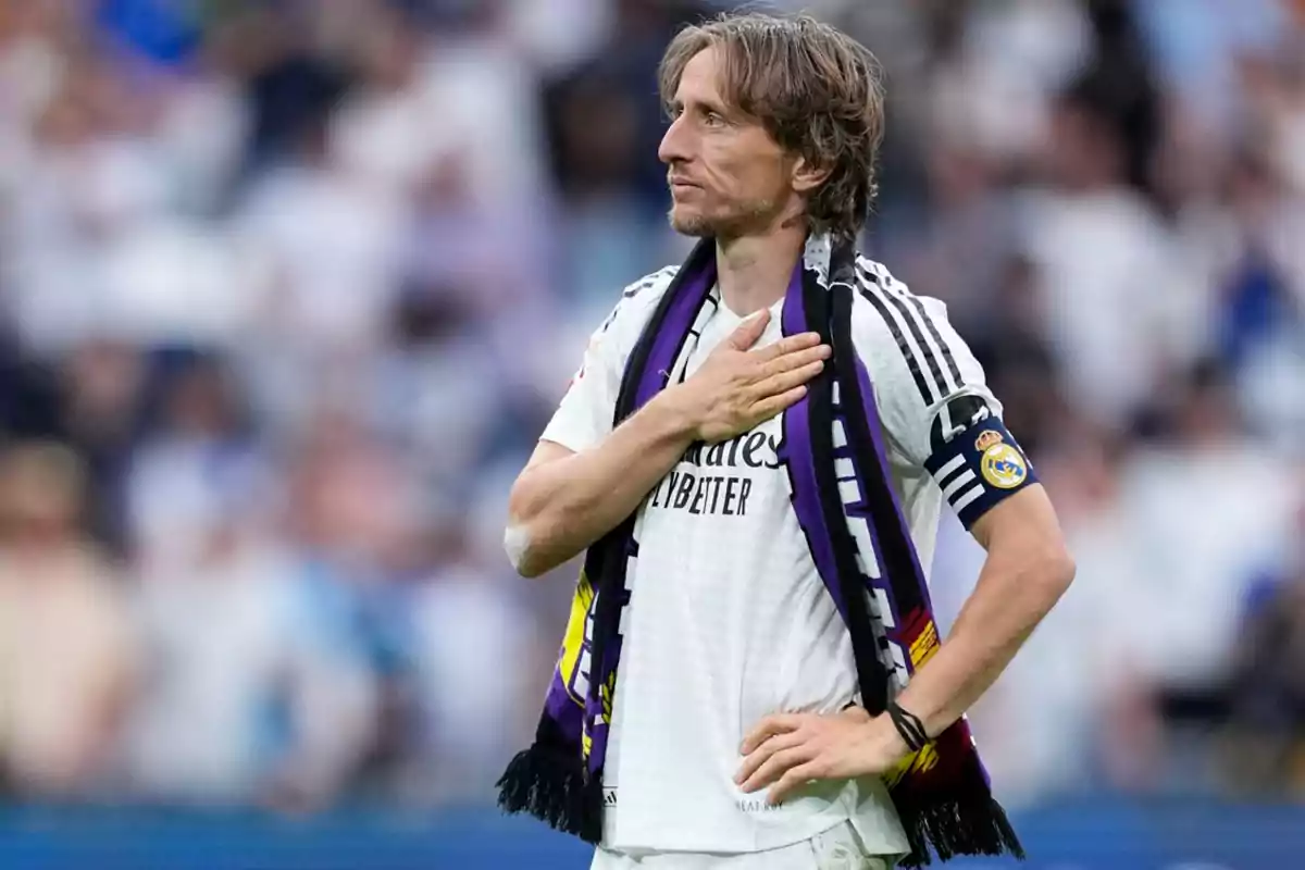 Un jugador de fútbol con uniforme blanco y bufanda en el cuello se lleva la mano al pecho en un estadio lleno de espectadores. Un jugador de fútbol con uniforme blanco y bufanda en el cuello se lleva la mano al pecho en un estadio lleno de espectadores.