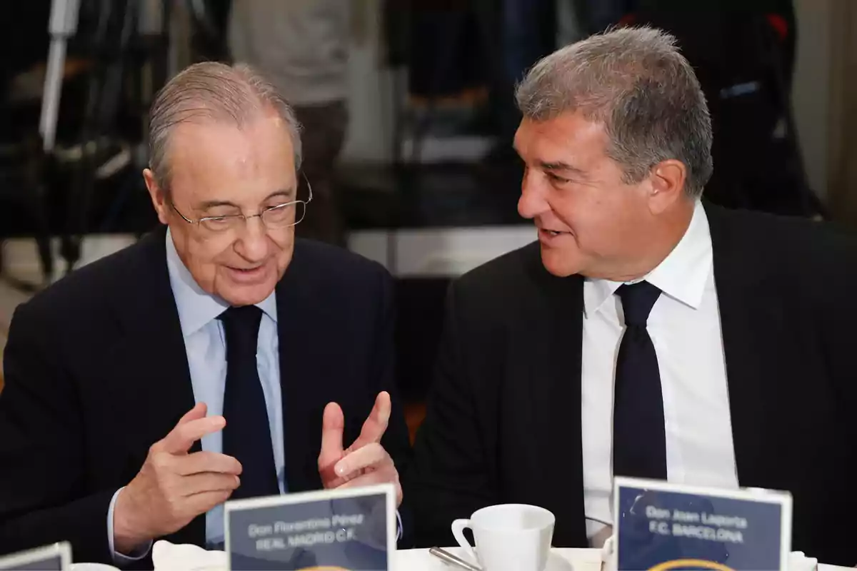 Two men in suits are sitting at a table talking during a formal event.