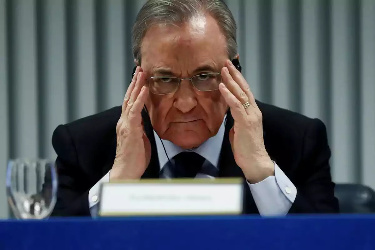 Hombre mayor con gafas y auriculares sentado en una mesa azul, tocándose la cabeza con ambas manos y mirando al frente con expresión seria