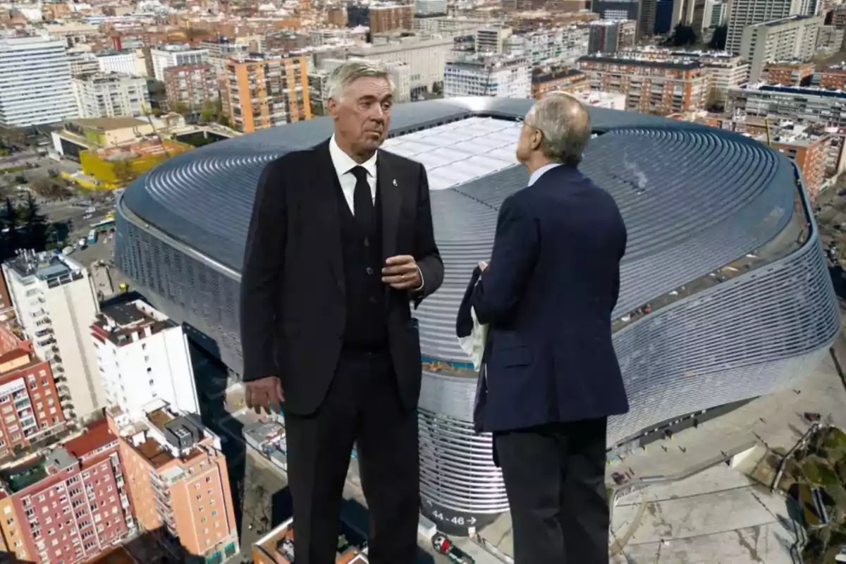 Two men stand talking in front of a modern stadium in a city full of buildings.