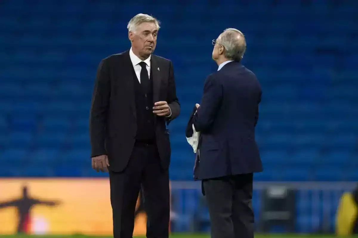 Dos hombres de traje conversan en un estadio vacío.