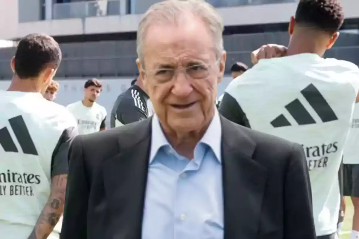 Hombre mayor con gafas y traje en primer plano mientras al fondo varios jugadores de fútbol entrenan con camisetas blancas