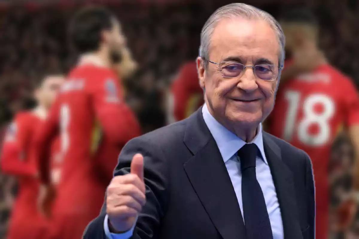 An older man wearing glasses and a suit smiles as he gives a thumbs-up gesture, with soccer players out of focus in the background.