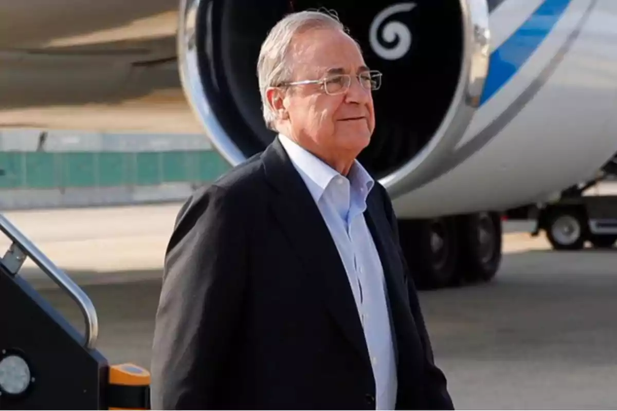 A man in a dark suit and light jersey is standing in front of a plane at an airport.