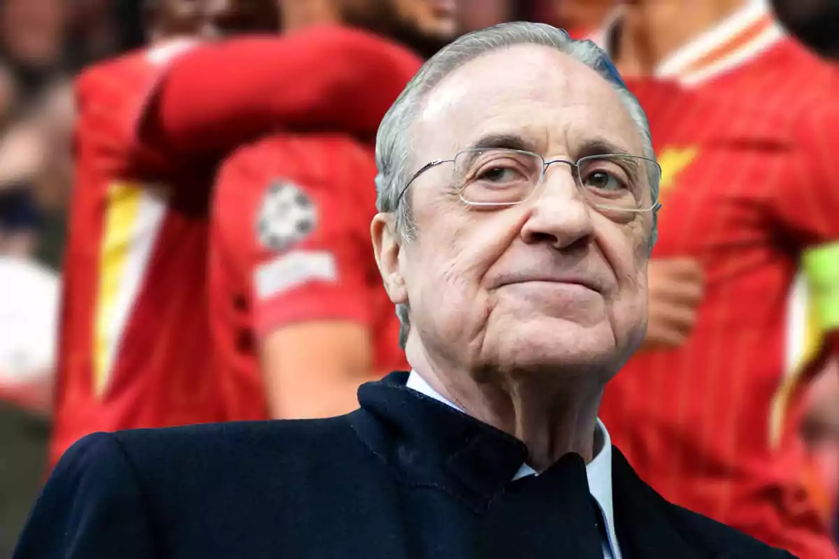 An older man with glasses and a dark suit in the foreground with soccer players in red uniforms celebrating in the background.