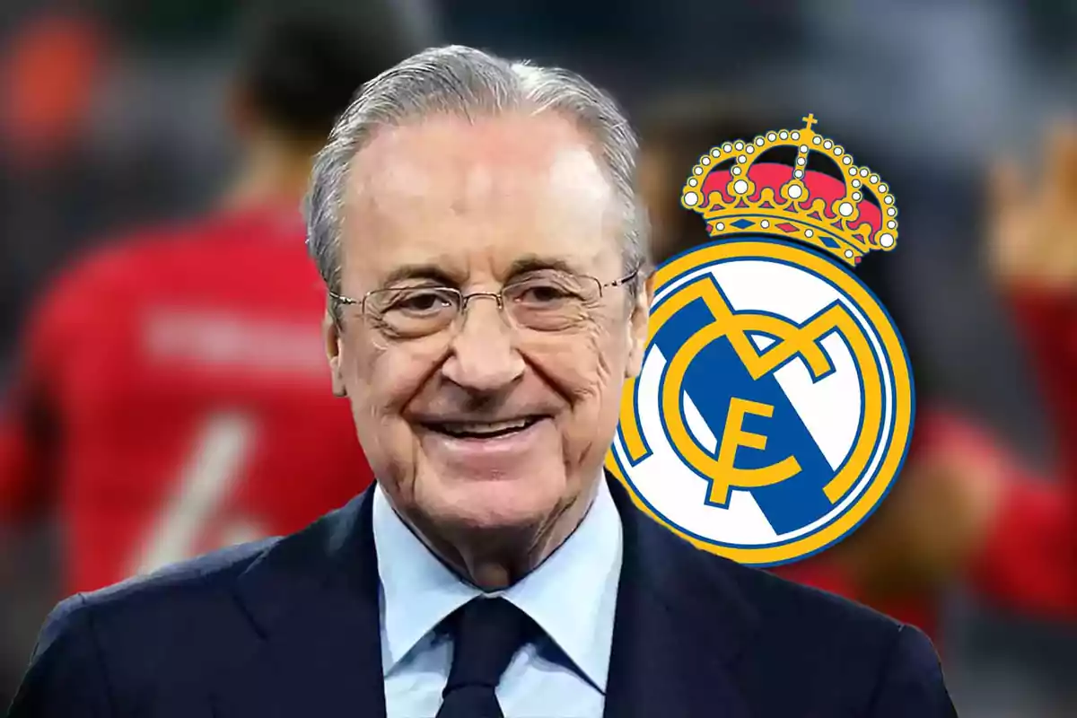 Un hombre mayor con gafas y traje sonríe frente al escudo del Real Madrid.