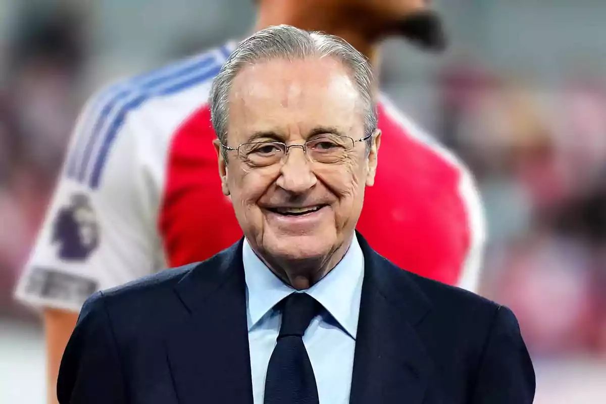 An older man wearing glasses and a suit smiles in front of an out-of-focus background of a soccer player.