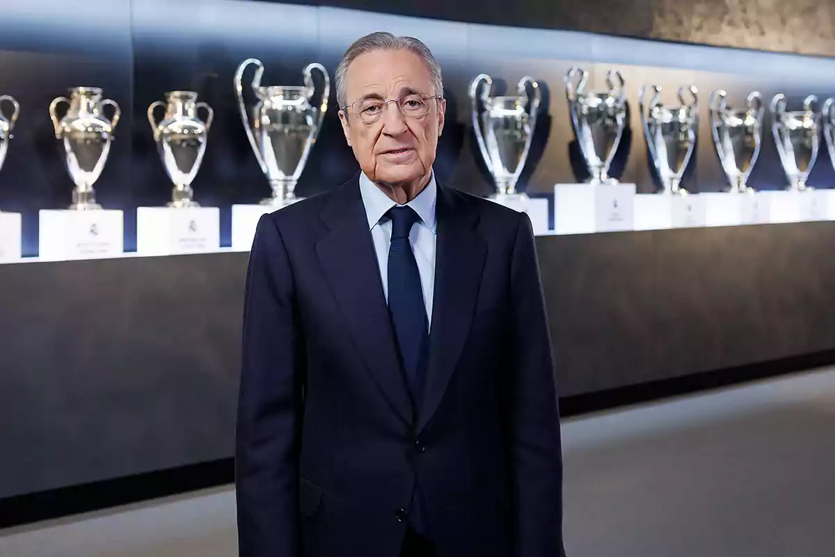 A man in a dark suit stands in front of a row of soccer trophies in a display case. A man in a dark suit stands in front of a row of soccer trophies in a display case.