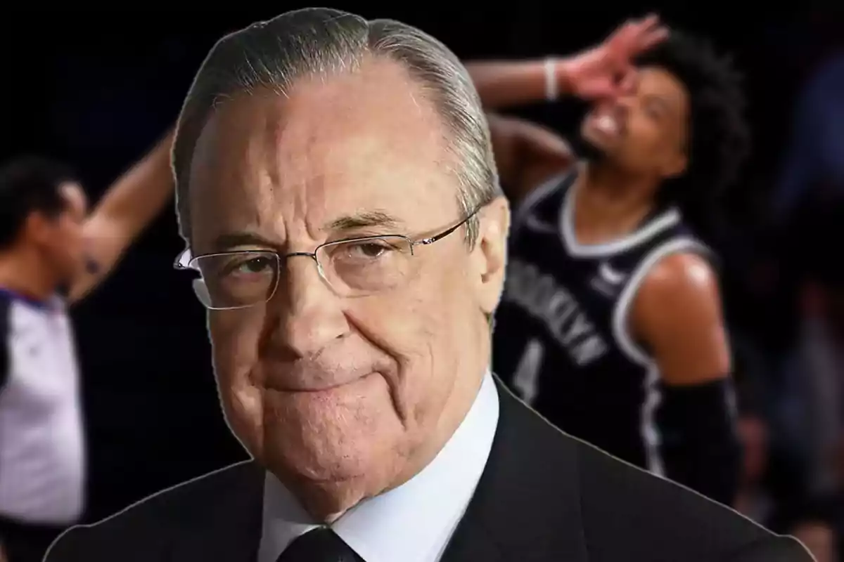 An older man wearing glasses and a suit smiles slightly as a basketball player can be seen celebrating in the background.