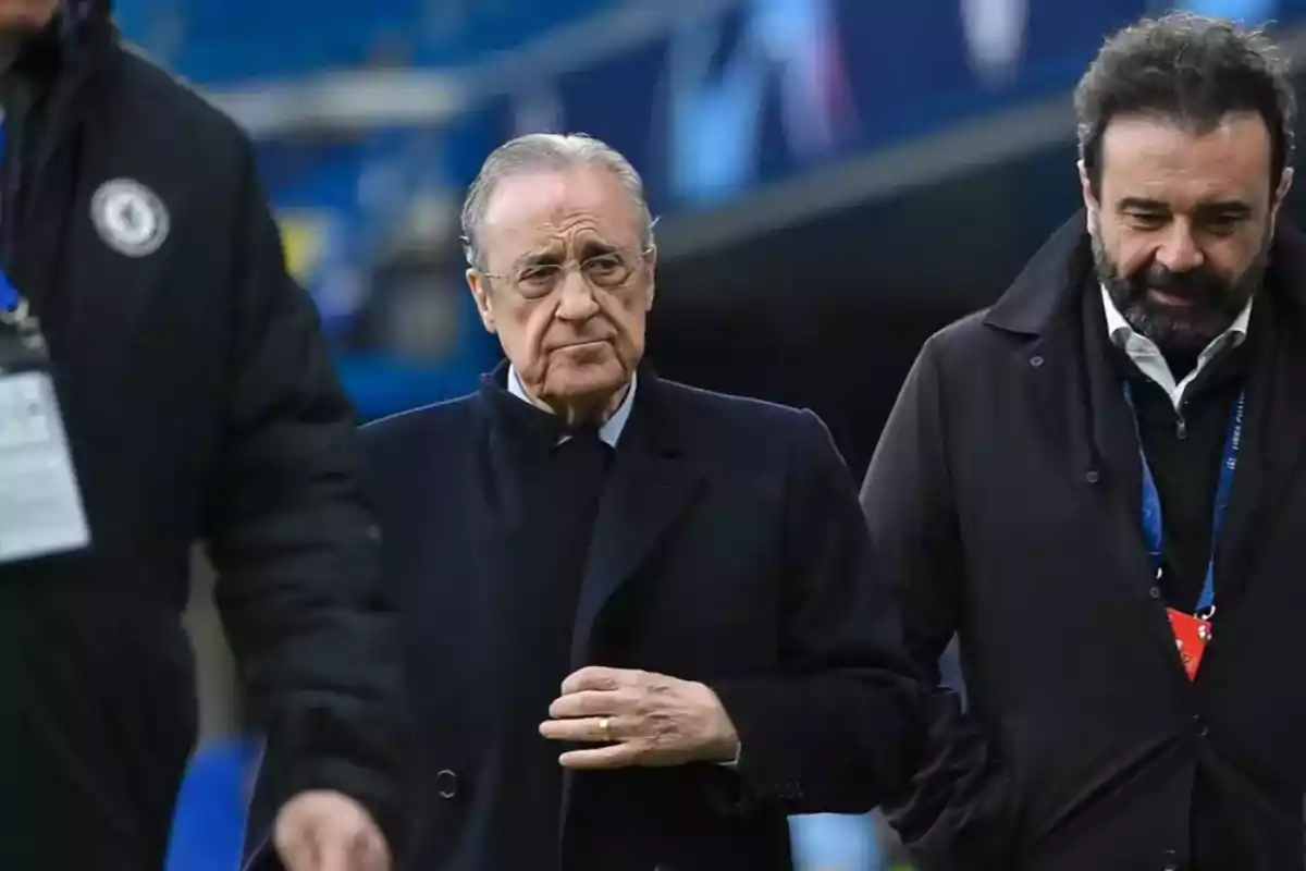 Un hombre mayor con gafas y abrigo oscuro camina junto a otro hombre con barba en un entorno deportivo.