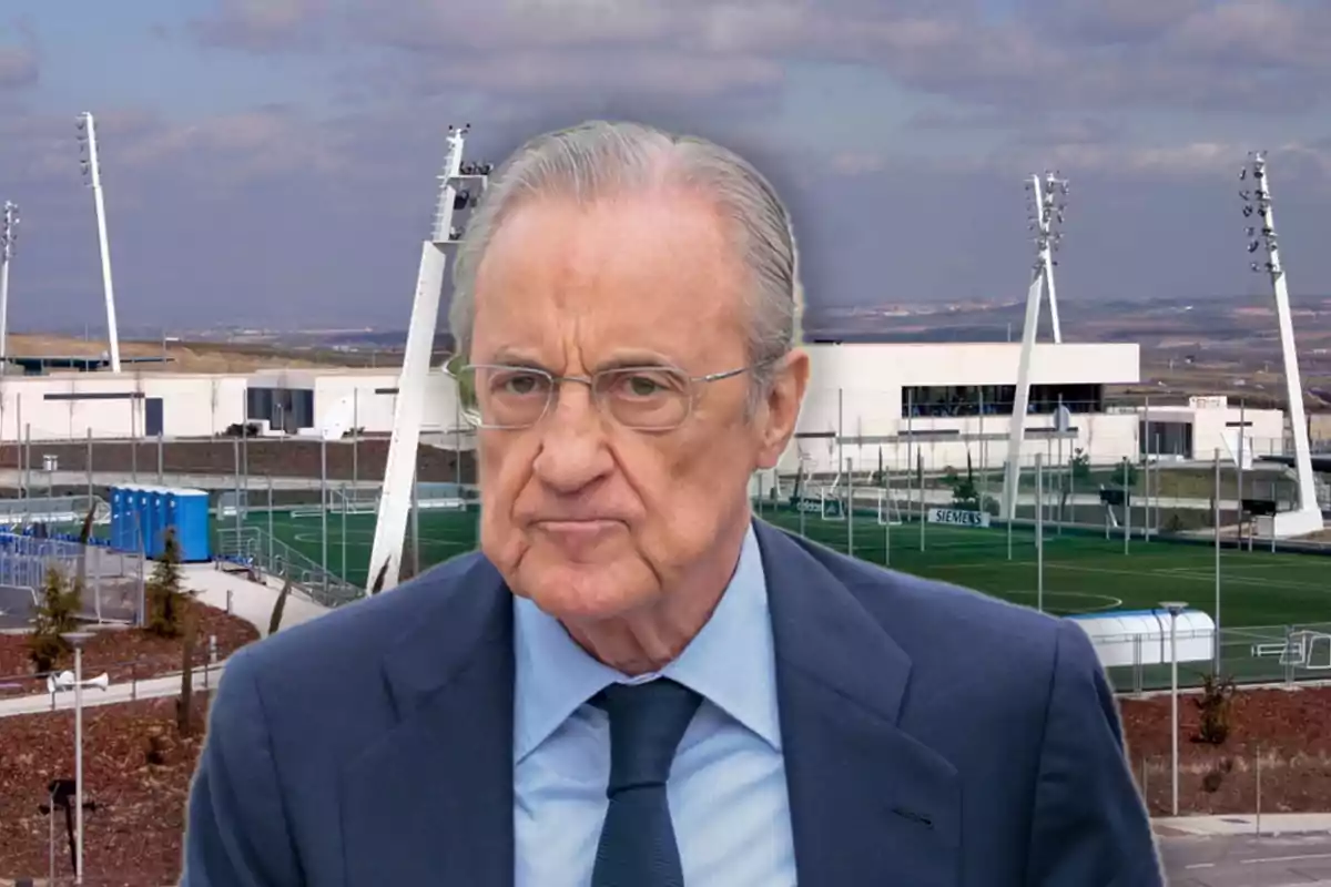 An older man with glasses and a dark suit is in front of a sports complex with soccer fields and lighting towers.
