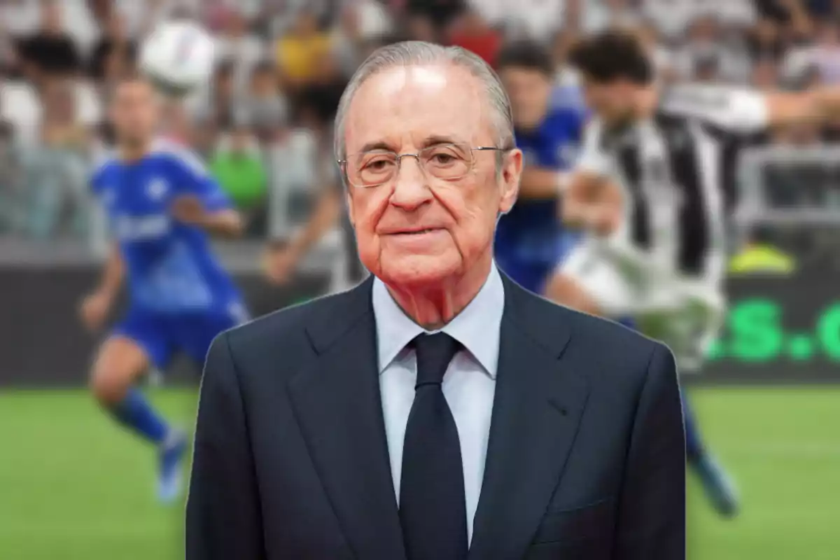 Older man with glasses and a dark suit posing in front of a soccer field with blurred players in the background