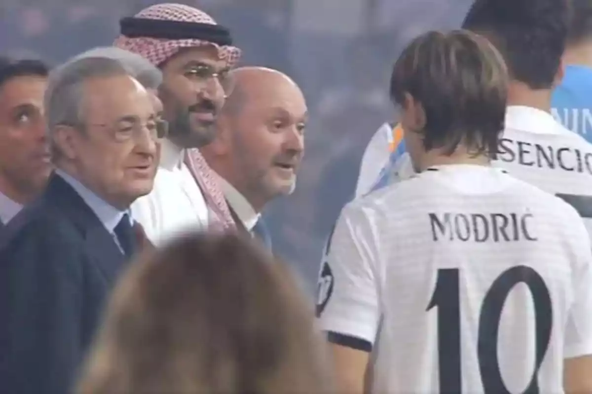 Un grupo de personas conversando en un evento deportivo, con un jugador de fútbol de espaldas.