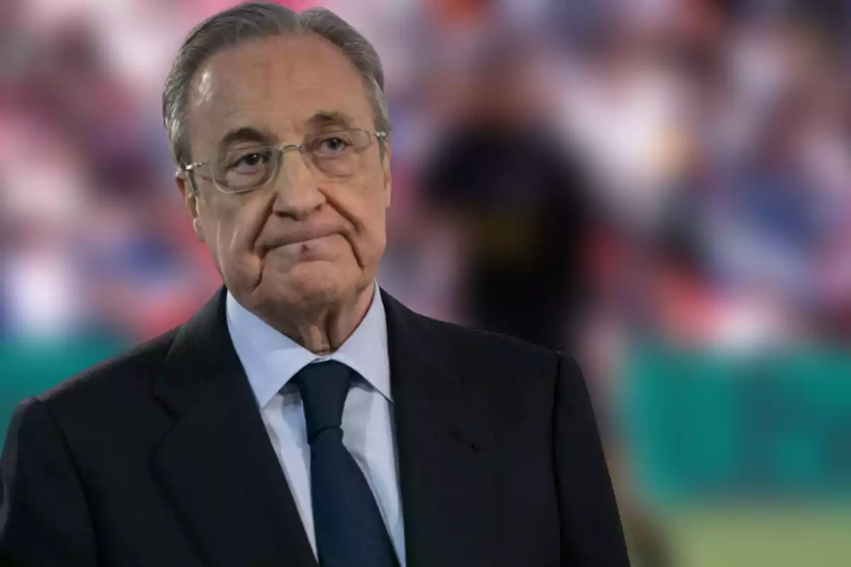 Un hombre mayor con gafas y traje oscuro está de pie frente a un fondo desenfocado.