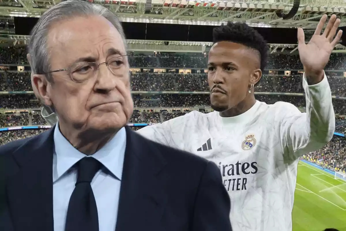 Un hombre con traje y gafas está en primer plano mientras un jugador de fútbol con uniforme blanco levanta las manos en el fondo de un estadio lleno de espectadores.