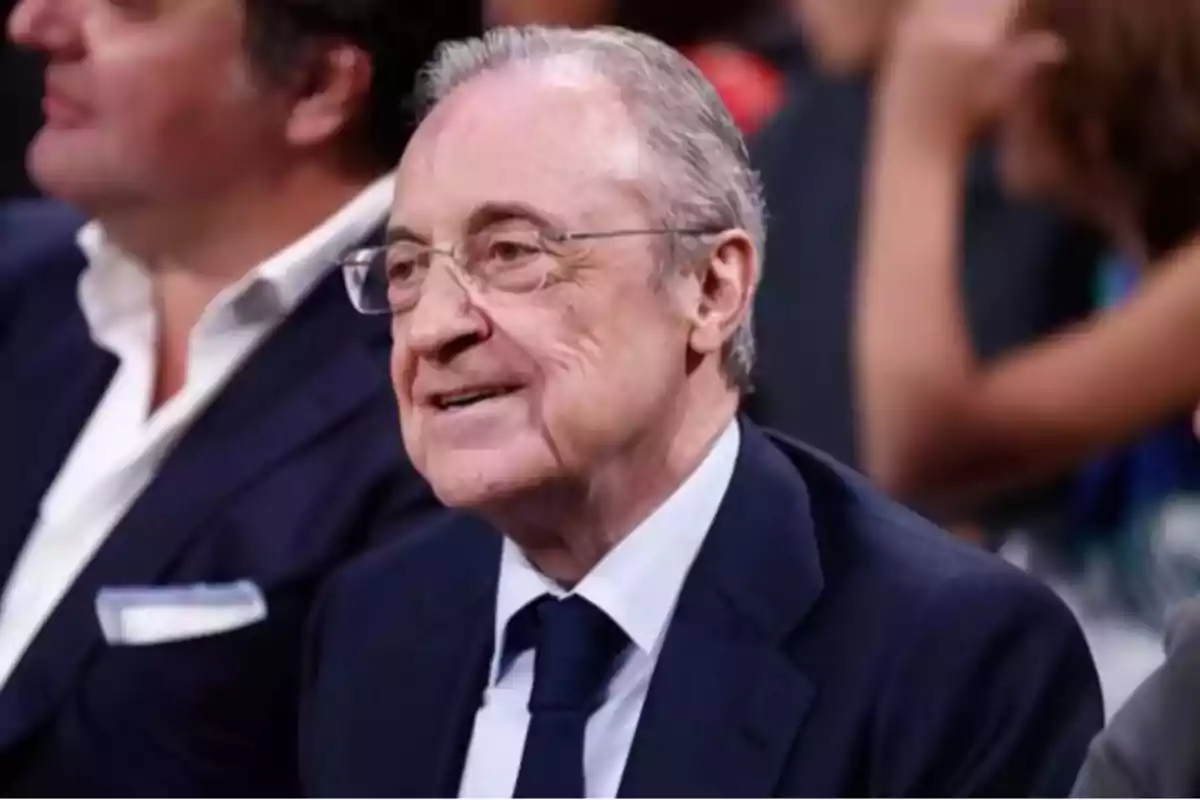 Un hombre mayor con gafas y traje oscuro sonríe mientras está sentado en un evento. Un hombre mayor con gafas y traje oscuro sonríe mientras está sentado en un evento.