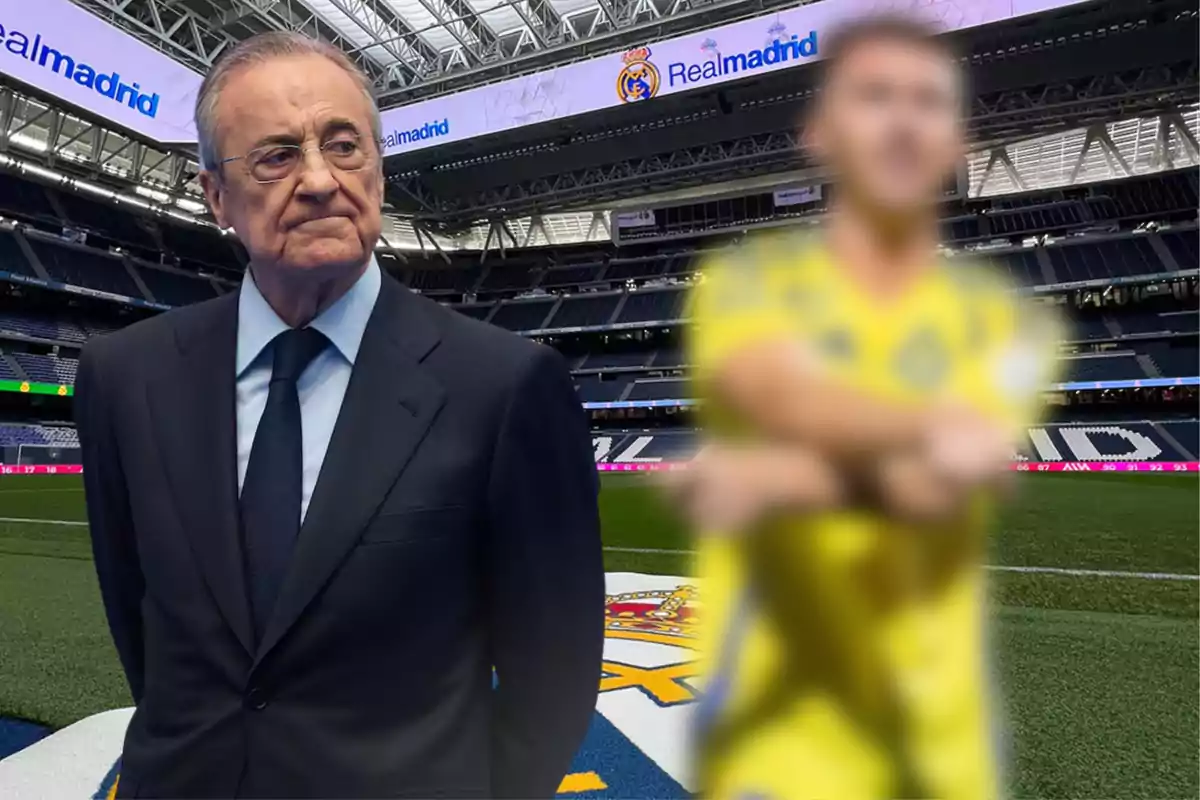 Un hombre de traje oscuro está de pie en un estadio de fútbol con el logo de Real Madrid en el césped y una figura desenfocada con uniforme amarillo a su lado