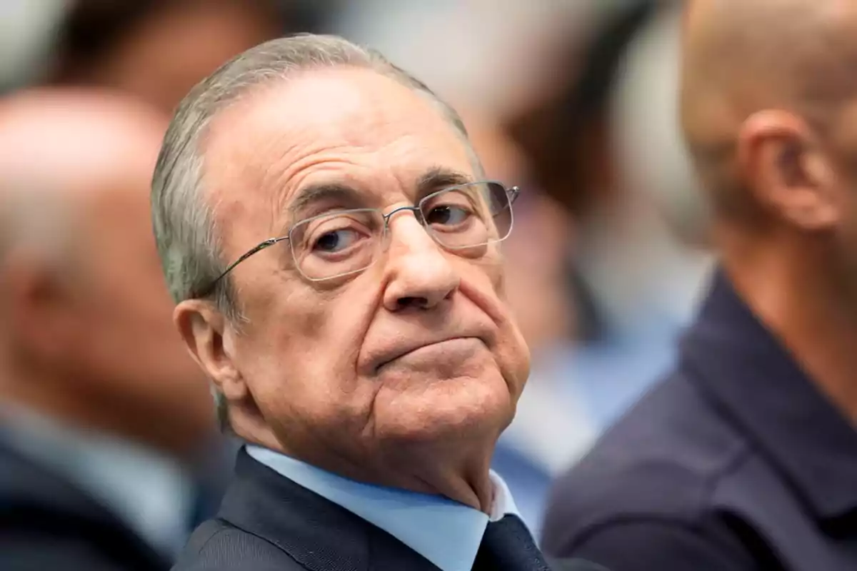 Un hombre mayor con gafas y traje oscuro mira hacia un lado en un entorno formal. Un hombre mayor con gafas y traje oscuro mira hacia un lado en un entorno formal.