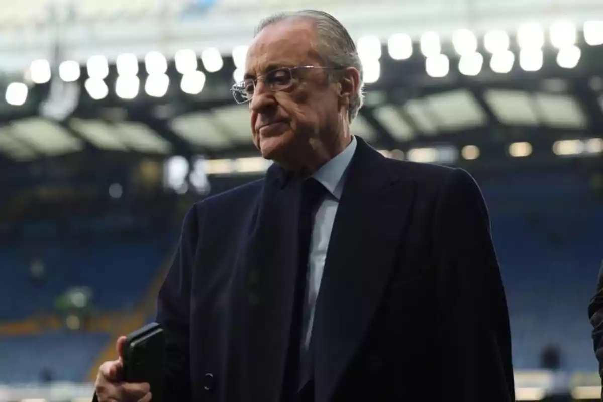 An older man wearing glasses and a dark suit holds a phone in a brightly lit stadium. An older man wearing glasses and a dark suit holds a phone in a brightly lit stadium.