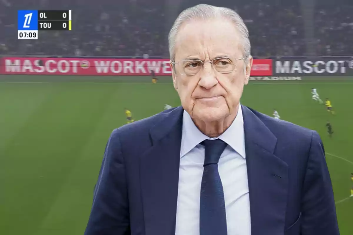 A man in a suit and tie is standing in front of a soccer field with a scoreboard in the upper left corner.