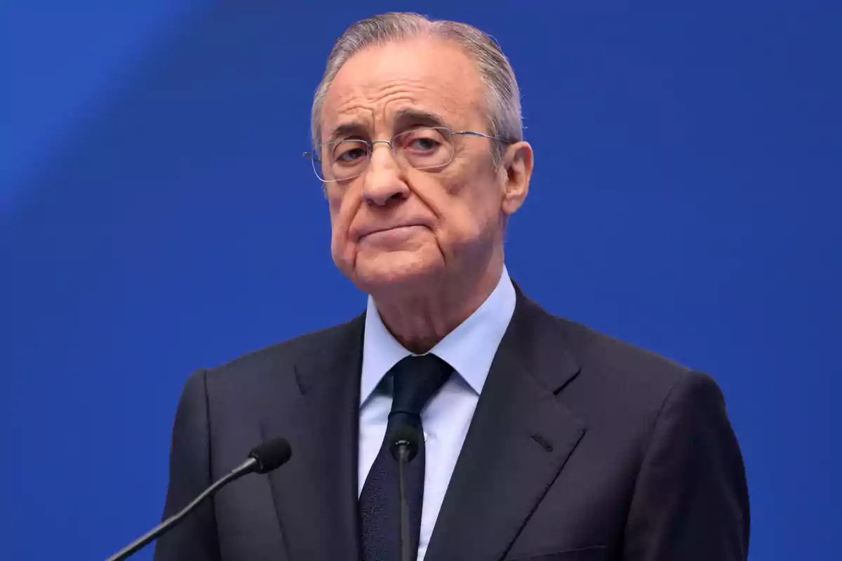 An older man with glasses and a dark suit in front of a blue background. An older man with glasses and a dark suit in front of a blue background.