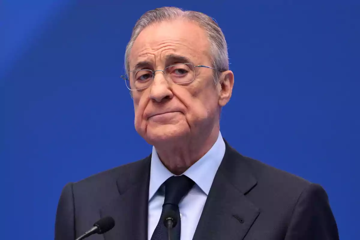 An older man with glasses and a dark suit in front of a blue background. An older man with glasses and a dark suit in front of a blue background.