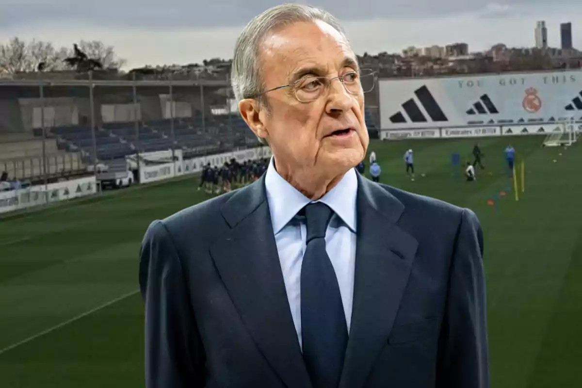 Un hombre con traje y corbata está de pie frente a un campo de entrenamiento de fútbol.