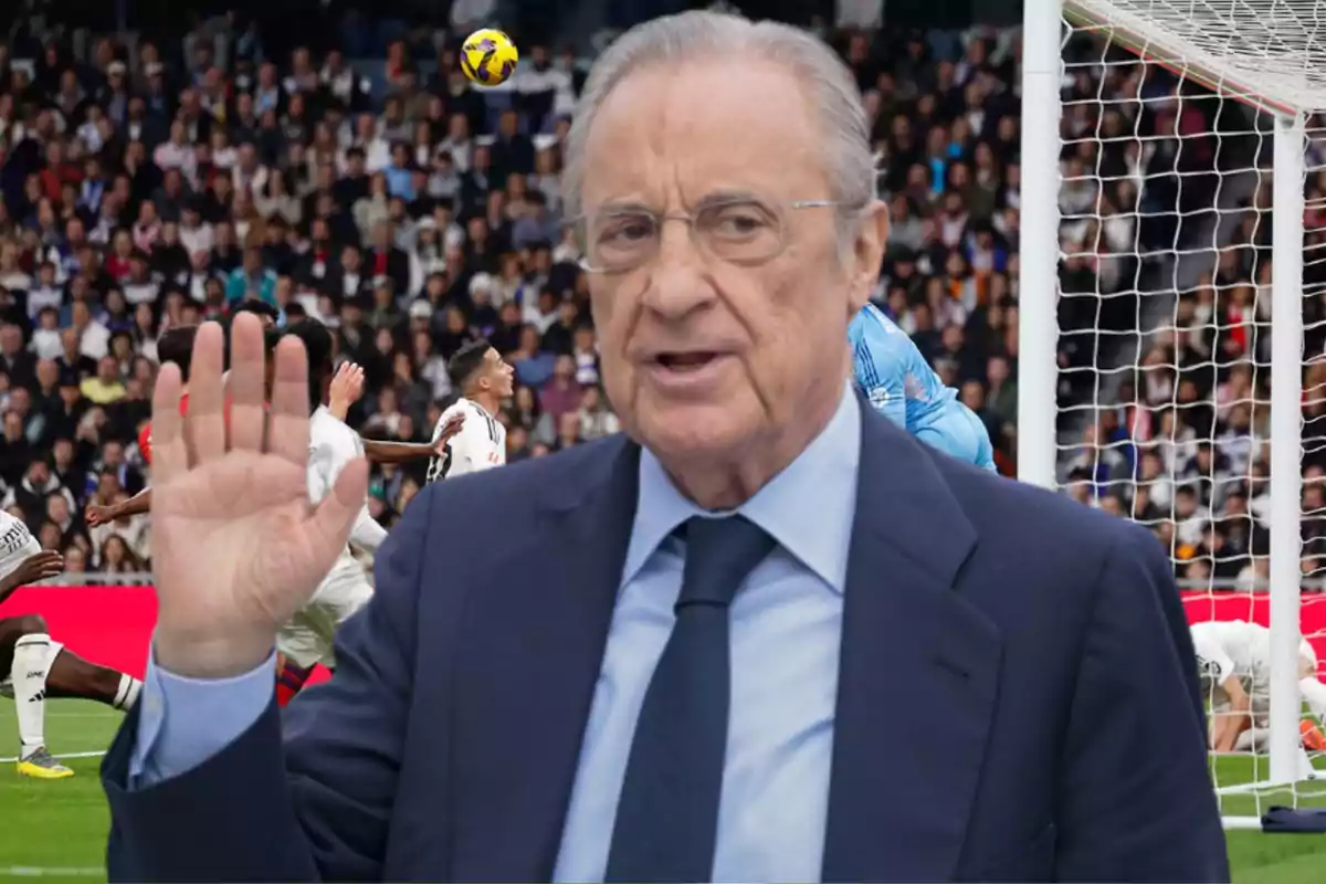 A man in a suit and tie raises his hand in front of a stadium full of spectators as a soccer match is being played.