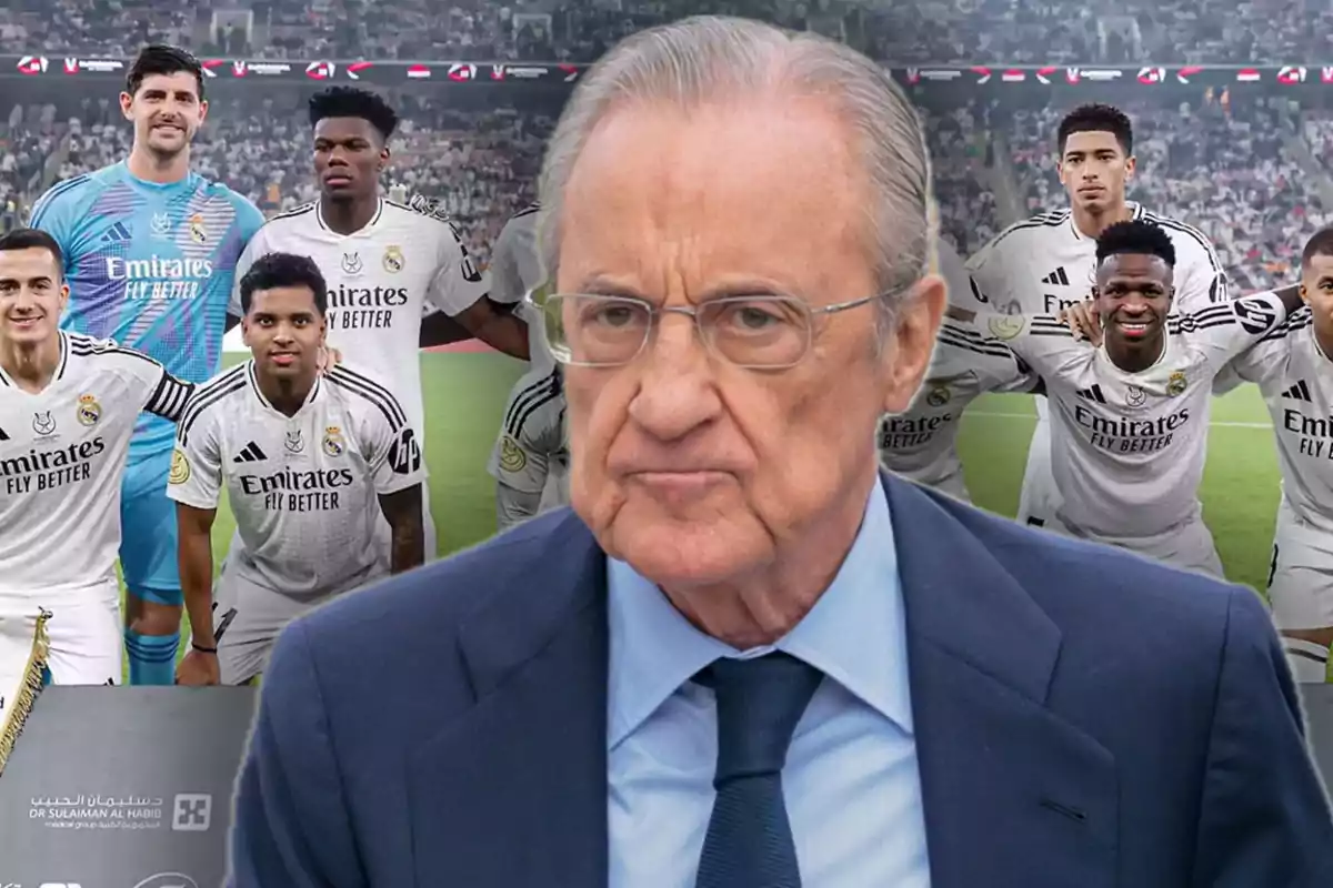 An older man with glasses and a suit stands in the foreground while a group of soccer players in white uniforms stand in the background in a stadium.