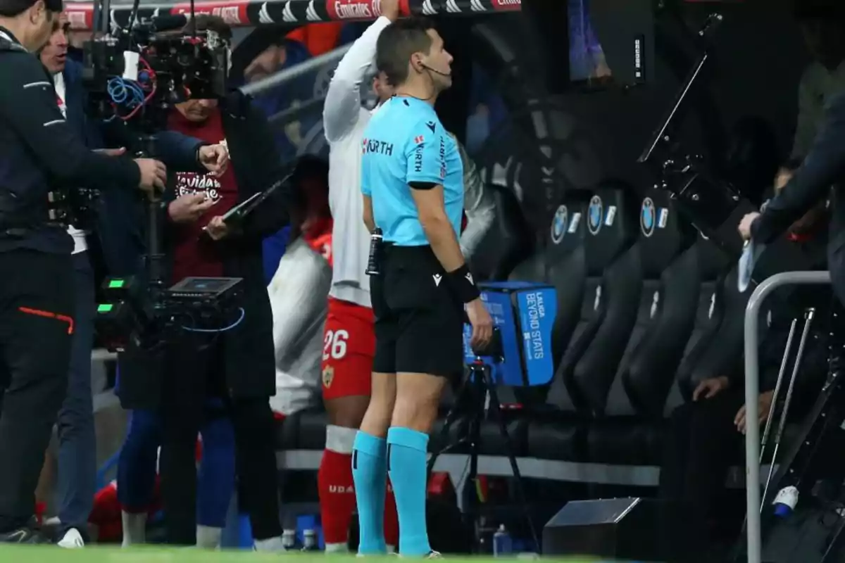 A soccer referee checks a screen in the technical area as several people surround him, including a cameraman and a player wearing the number 26. A soccer referee checks a screen in the technical area as several people surround him, including a cameraman and a player wearing the number 26.