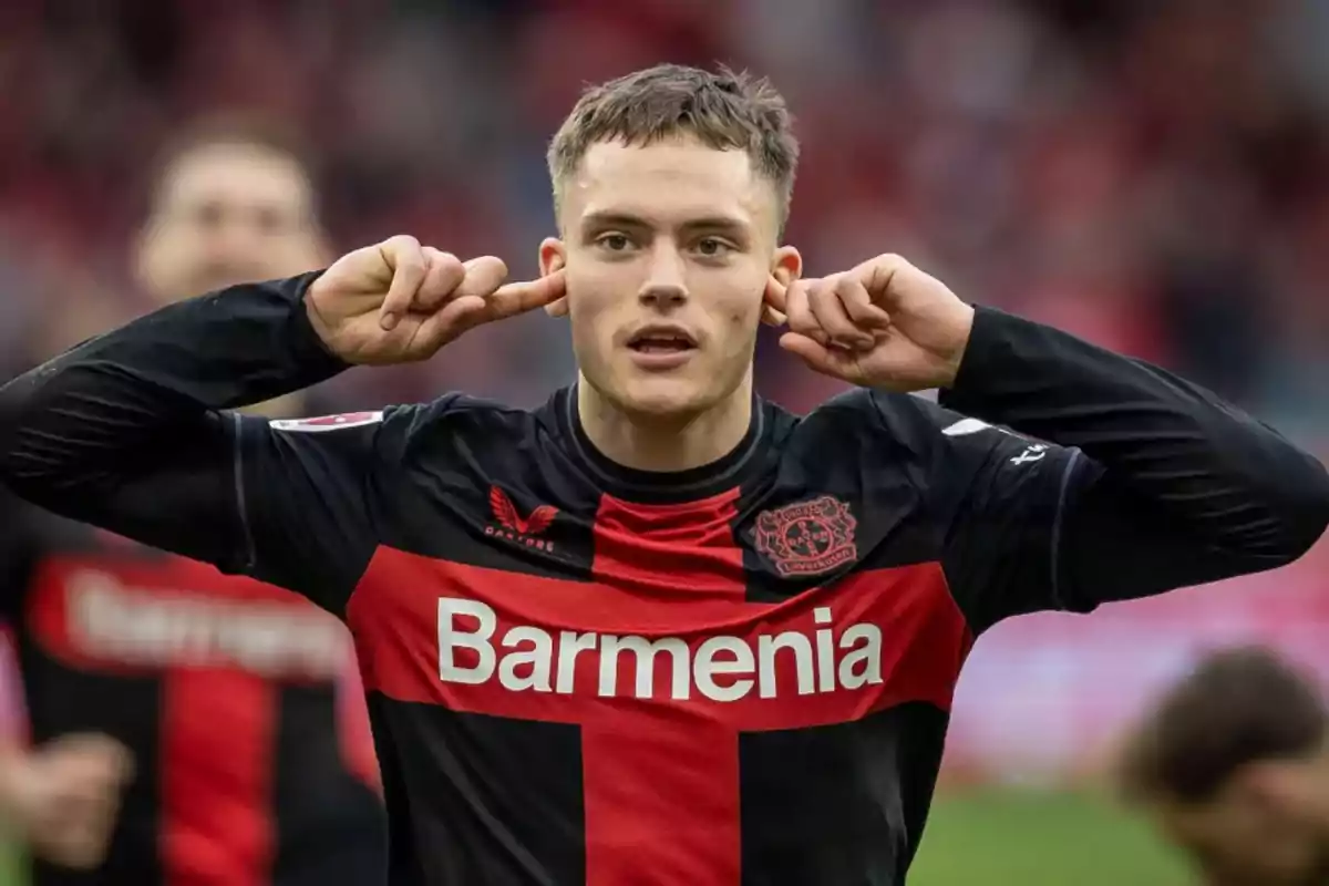 Un jugador de fútbol con uniforme negro y rojo se tapa los oídos con los dedos en un estadio.