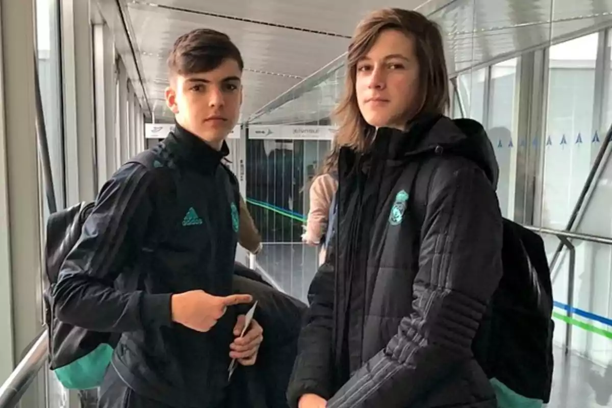 Dos jóvenes con ropa deportiva negra y mochilas posan en un pasillo acristalado. Dos jóvenes con ropa deportiva negra y mochilas posan en un pasillo acristalado.