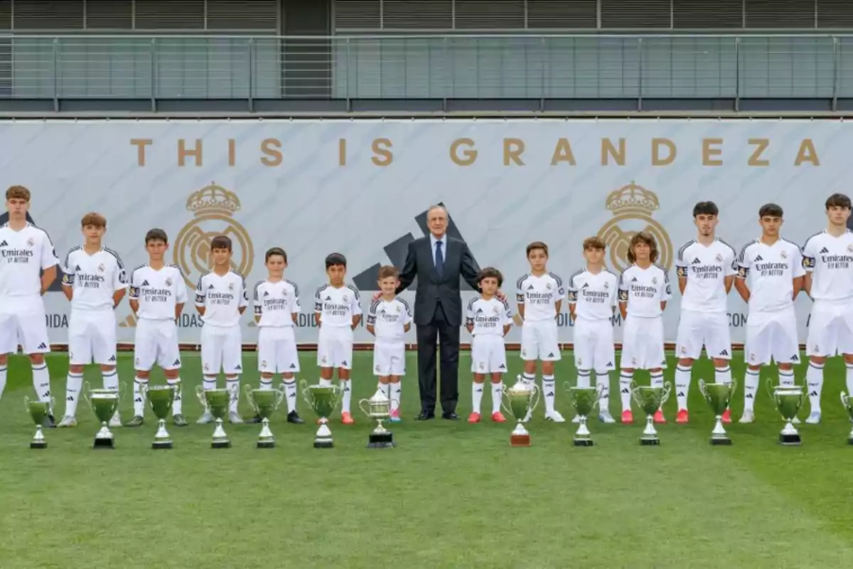 A group of young players in white uniforms and a man in a suit pose in a row in front of several trophies on the grass, with a background showing the Real Madrid crest and the phrase "This is grandeza."
