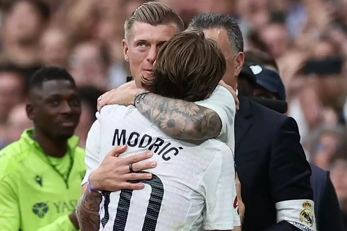 A player with the name "Modric" on his jersey hugs another player while a person in a green uniform watches in the background.
