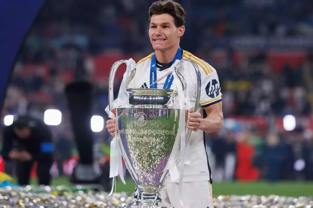 Un jugador de fútbol sonríe mientras sostiene un gran trofeo en un estadio lleno de confeti. Un jugador de fútbol sonríe mientras sostiene un gran trofeo en un estadio lleno de confeti.
