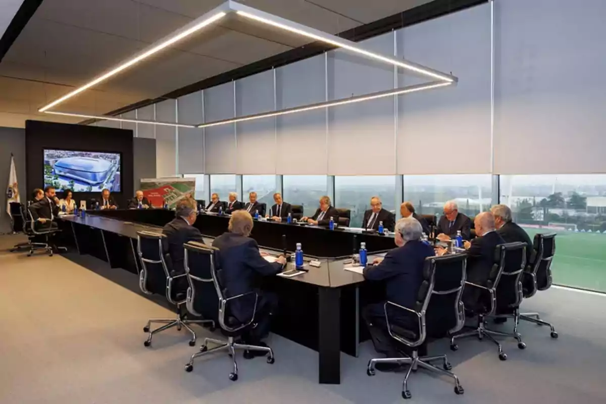 A group of people in a modern conference room with a large rectangular table and screens showing images of a stadium. A group of people in a modern conference room with a large rectangular table and screens showing images of a stadium.