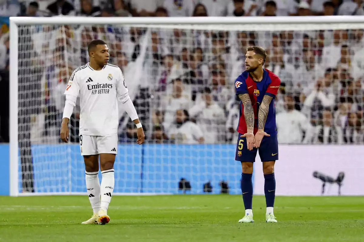 Two footballers on the field during a match, one in a white uniform and the other in a blue and red uniform, with a crowd of spectators in the background.