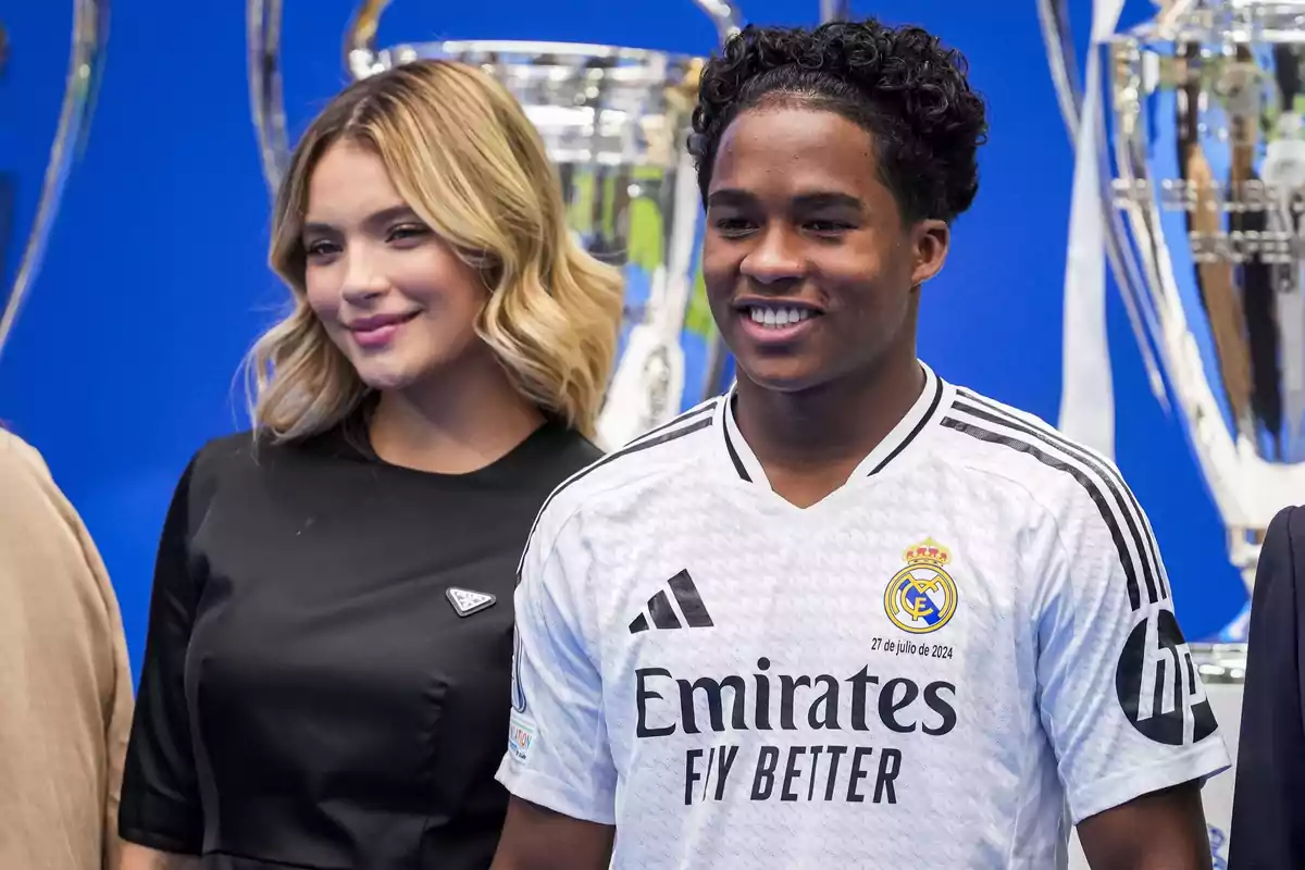 A woman and a man smile in front of trophies, he is wearing a Real Madrid soccer jersey.