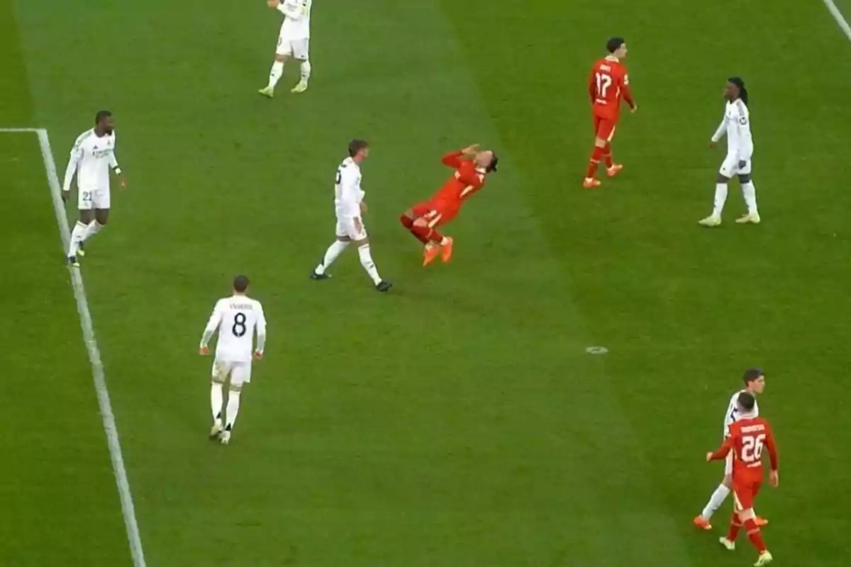 Jugadores de fútbol en el campo durante un partido, uno de ellos en uniforme rojo parece estar cayendo hacia atrás mientras otros jugadores en uniforme blanco observan. Jugadores de fútbol en el campo durante un partido, uno de ellos en uniforme rojo parece estar cayendo hacia atrás mientras otros jugadores en uniforme blanco observan.
