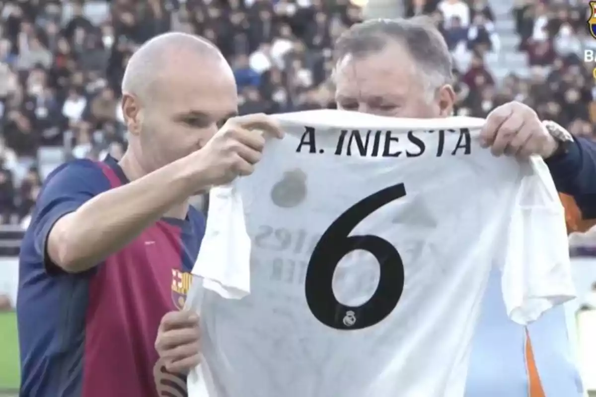 Dos personas sostienen una camiseta de fútbol con el nombre "A. Iniesta" y el número 6 en un estadio lleno de espectadores.