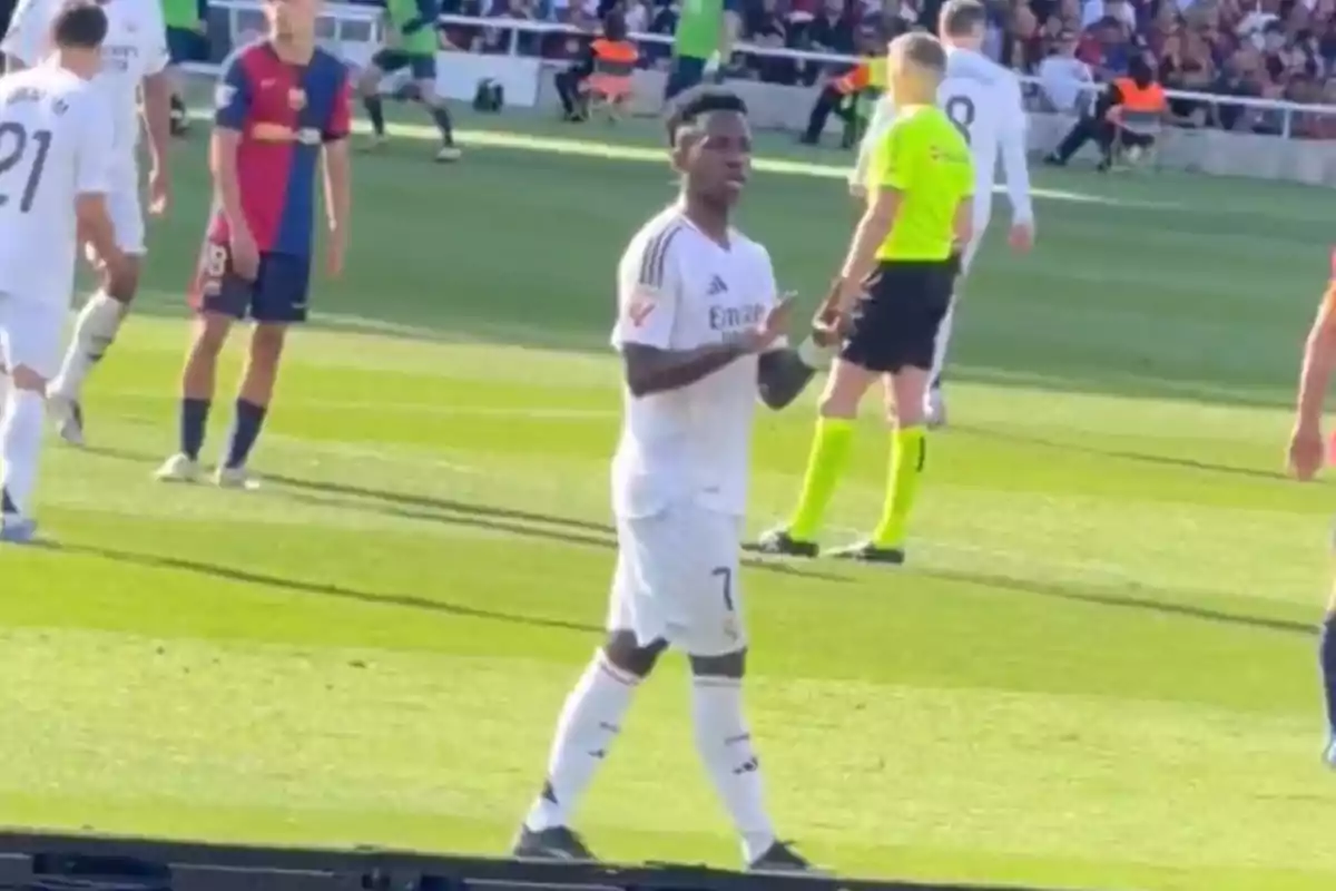 Un jugador de fútbol con uniforme blanco en el campo durante un partido, con otros jugadores y un árbitro cerca.
