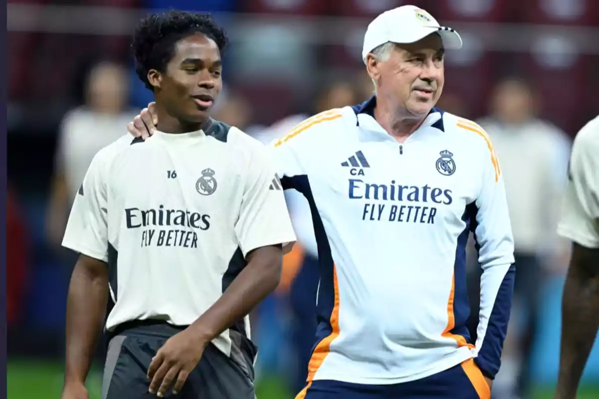 Dos personas con ropa de entrenamiento del Real Madrid conversan y sonríen en un campo de fútbol.