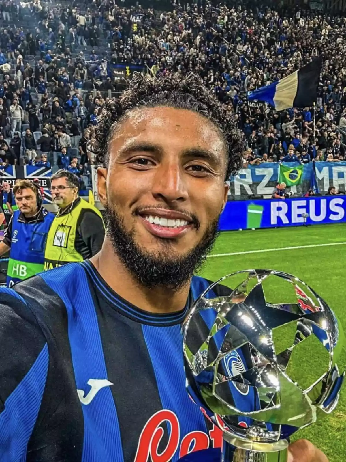 Un jugador de fútbol sonríe mientras sostiene un trofeo en el campo, con una multitud de aficionados en el fondo.