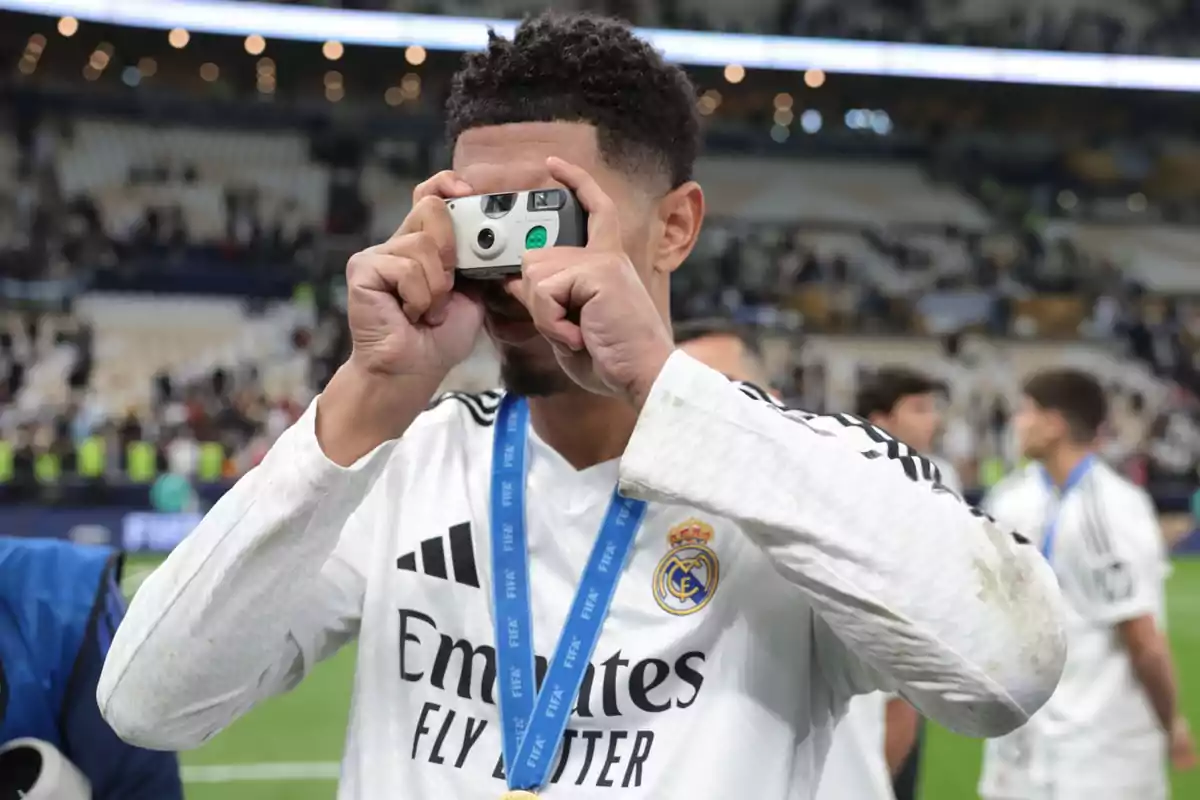 Un jugador de fútbol con uniforme blanco y medalla toma una foto con una cámara desechable en un estadio.